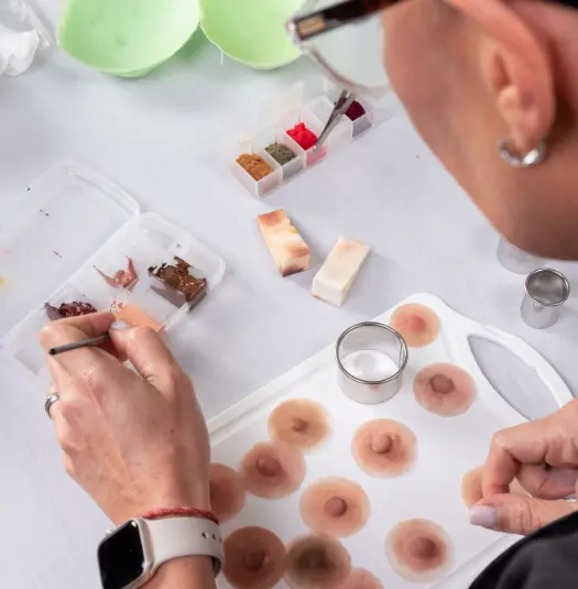 Una persona con gafas y un reloj inteligente está sentada en una mesa trabajando en unas pequeñas prótesis con forma de pezón. Sobre la mesa hay varios pigmentos, herramientas y otros materiales. La persona pinta y detalla meticulosamente la areola de las prótesis con un pincel.