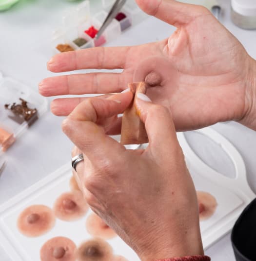 Una persona sosteniendo una prótesis de pezón de silicona con múltiples pezones protésicos en una bandeja al fondo.