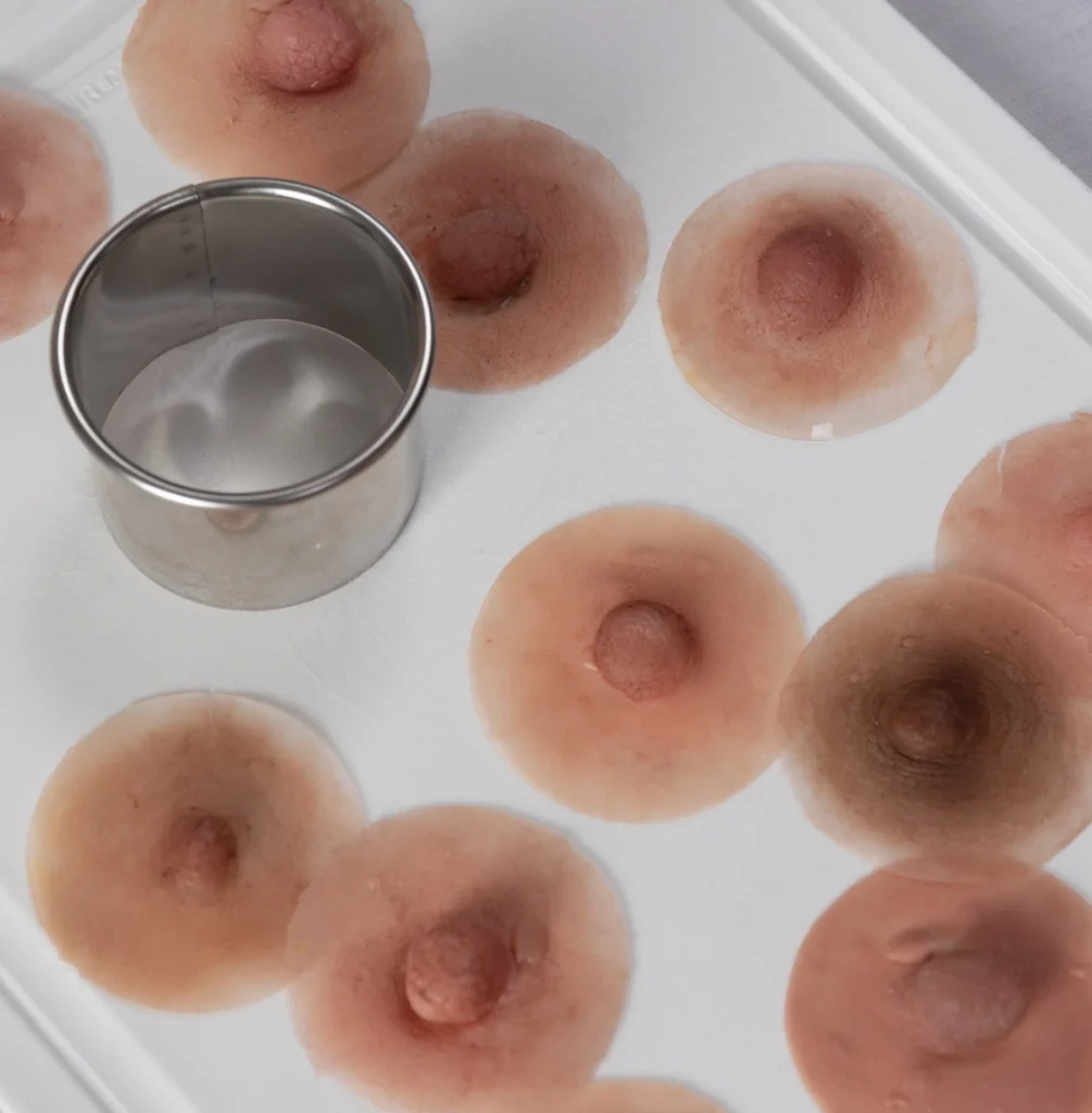 Una bandeja blanca con varias prótesis areolas de silicona realistas dispuestas alrededor de un cortador redondo de metal. Las prótesis están detalladas con distintos tonos de rosa y marrón, asemejándose a los pezones humanos naturales. El cortador se coloca a la izquierda de la bandeja.