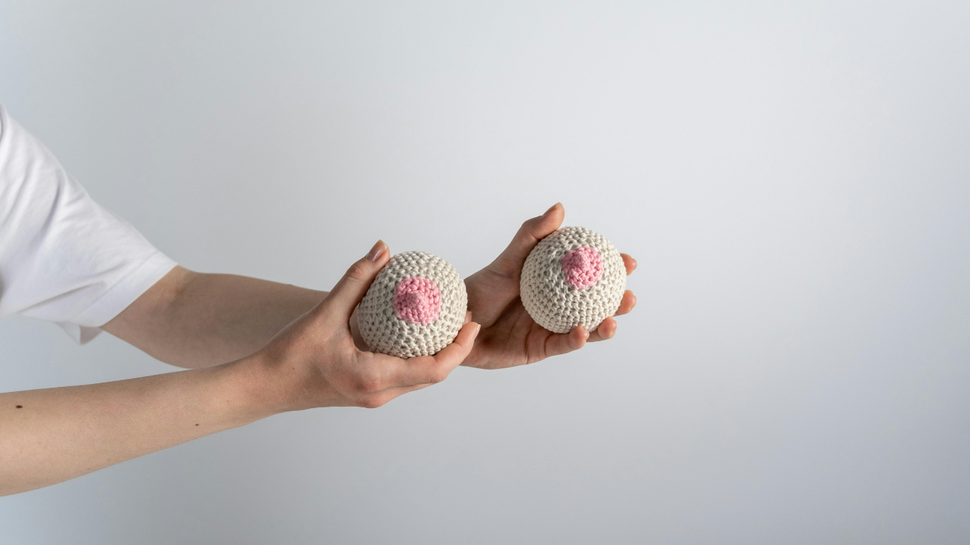 Reconstrucción mamaria: manos sostienen dos prótesis tejidas que simulan senos con pezones rosados, sobre fondo neutro.
