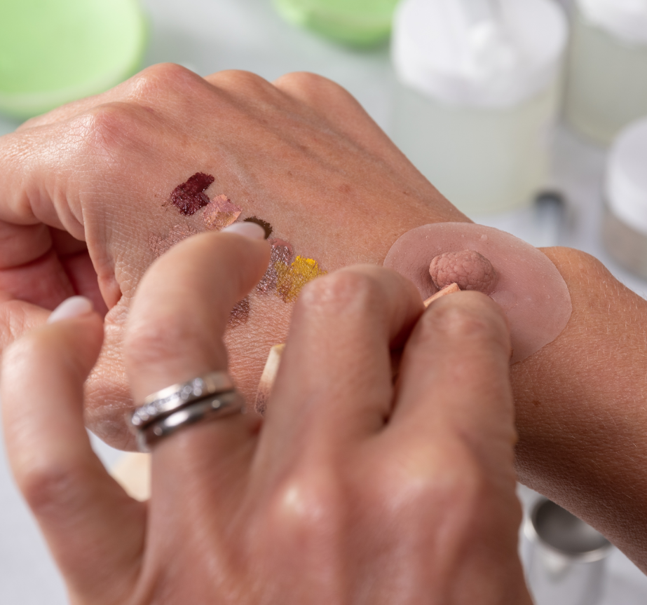 Una persona está probando productos de maquillaje cosmético en el dorso de su mano. Se ven varias muestras de diferentes colores y la persona está usando un pequeño aplicador para probar una muestra. Llevan un anillo de plata en su mano izquierda, tal vez explorando opciones para una prótesis de areola con tonos naturales.